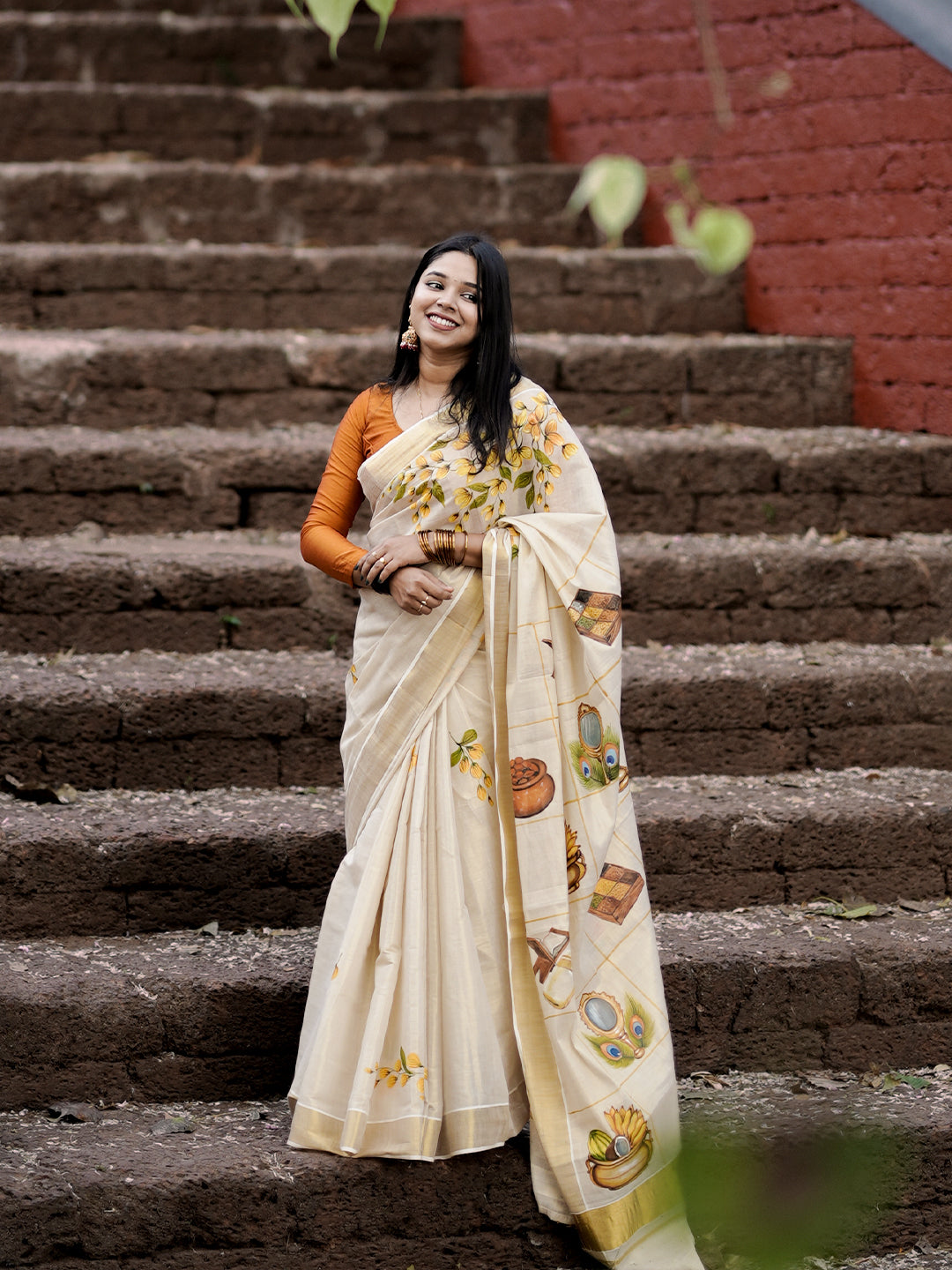 Woman in a white saree with gold patterns standing on stone steps.
