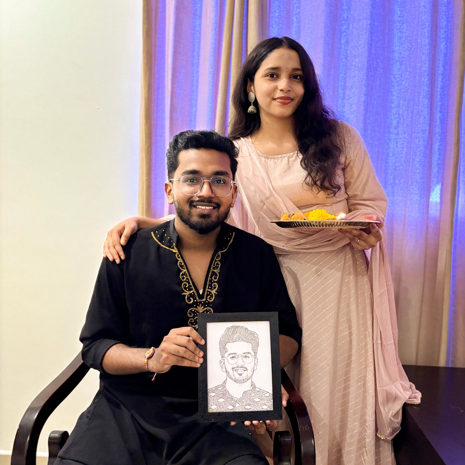 Man holding a framed portrait of a man, woman in traditional attire holding a plate, against a curtain background