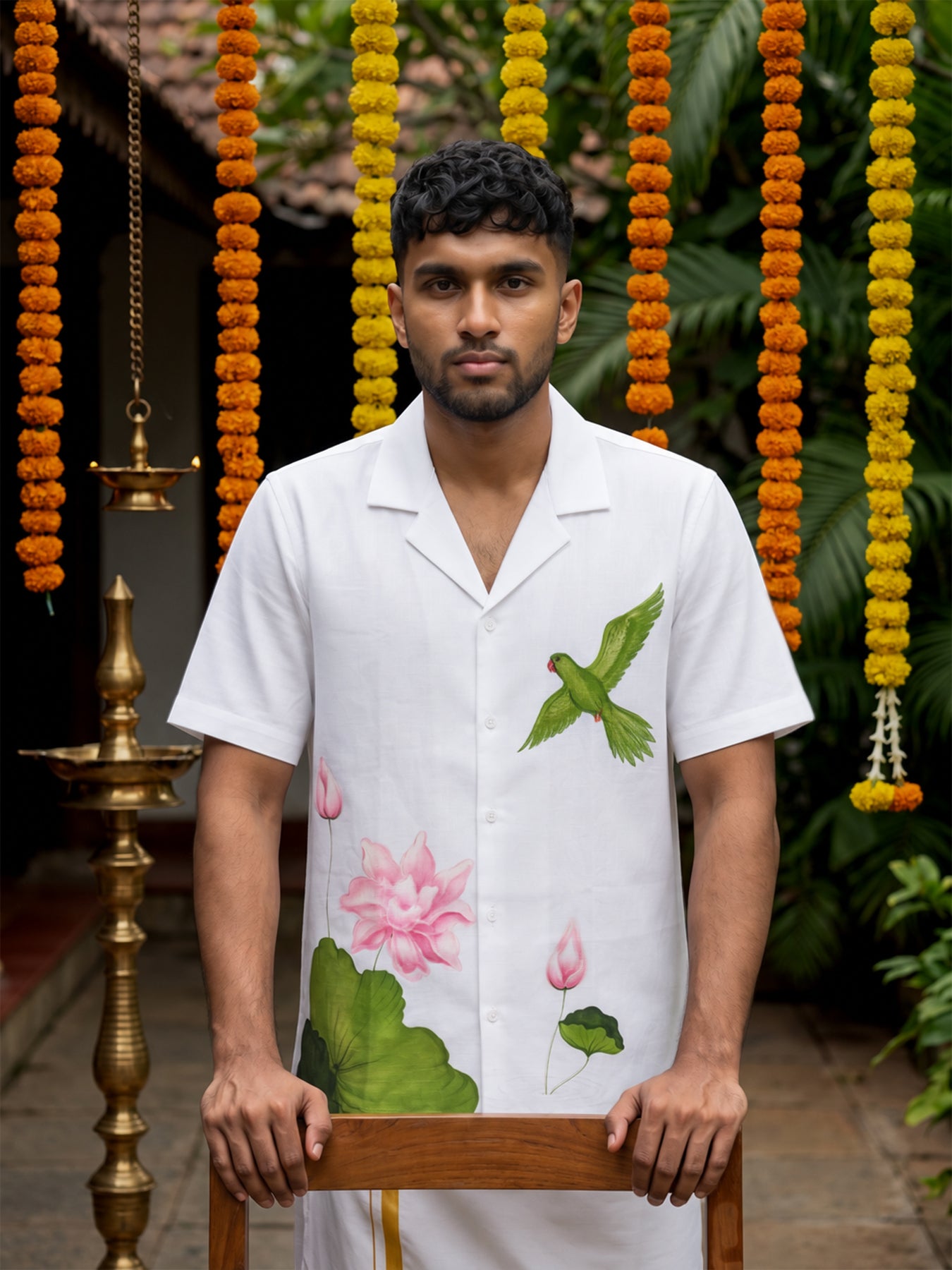 A man wearing a white short-sleeve button-up shirt with a green bird and pink lotus flower print, standing in front of a decorated background with hanging orange and yellow marigold garlands.